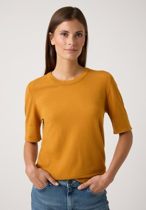 T-shirt jaune moutarde à manches courtes avec un col rond, en tissu tricoté. Présente une coupe décontractée et des ourlets roulés. Assorti avec un jean bleu clair.