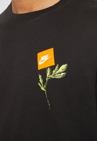Svart bomulls t-shirt med ett grönt bladvyk och en orange Nike-logotagg på bröstet. Slät textur med rund halsringning.