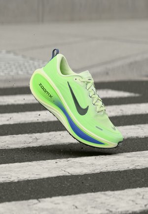 Neongrøn Nike ZoomX løbesko i luften over et fodgængerfelt på asfalt, der viser den sorte Swoosh og blå såldetalje.