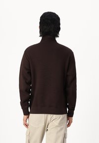 Sudadera marrón con cuello alto, mangas largas y puños acanalados. La tela parece suave y ligeramente texturada. Lleva puesta con pantalones cargo de color claro.
