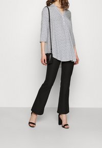 Kobieta ubrana w wzorzystą szaro-tunikę, czarne szerokie spodnie, czarne sandały na obcasie i nosząca małą czarną torbę na ramię.