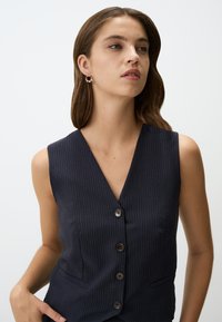 Gilet senza maniche navy con righe verticali, caratterizzato da un profondo scollo a V e cinque bottoni marroni. Sono visibili tasche su entrambi i lati.