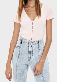 Kvinna iklädd en ljusrosa kortärmad ribbad topp med pärlknappar och högmidjade ljusblå utsvängda jeans med stora framfickor.