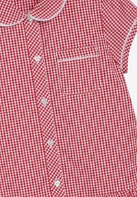 Rdeča in bela karo srajca s Peter Pan ovratnikom, kratkimi rokavi in prsnim žepom. Ima gumbe v obliki src s belim robom.