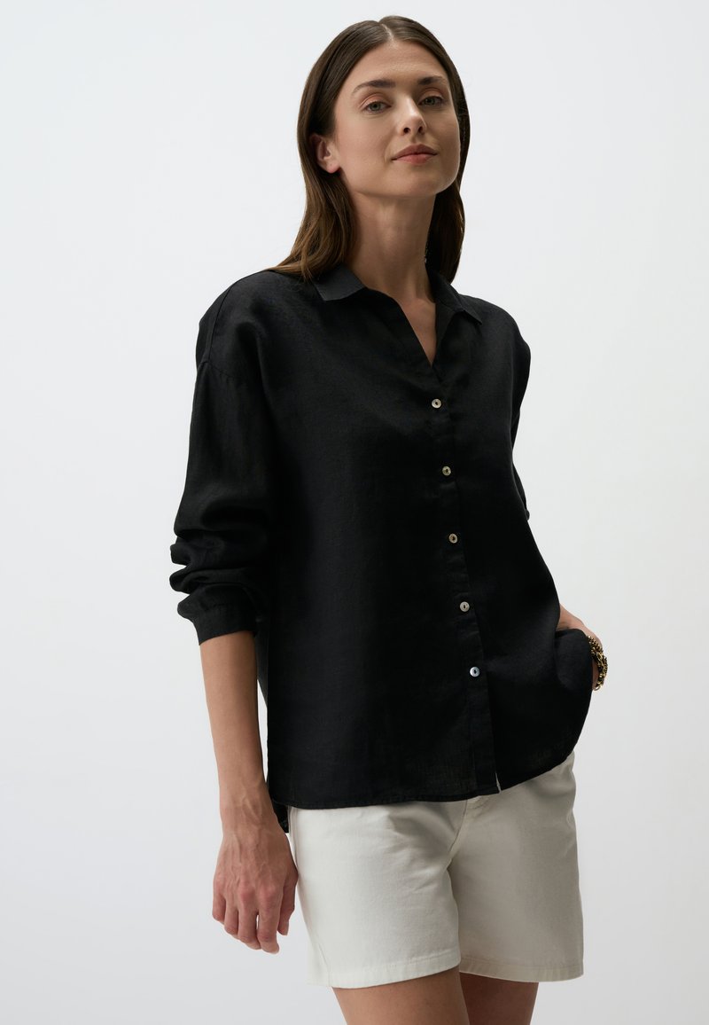 Camicia in lino nera con colletto, chiusura a bottoni sul davanti e maniche lunghe, abbinata a pantaloni corti bianchi. Texture liscia e vestibilità rilassata.