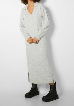 Personne portant une longue robe en maille gris clair à manches longues et col, associée à des bottines noires épaisses.