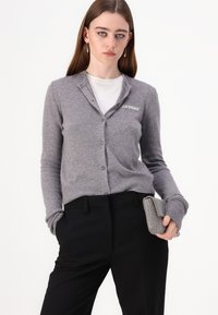 Grå cardigan med knaplukning fortil, ribbede manchetter og broderet "2ND DAY" på brystet, parret med sorte bukser og en ternet clutch.
