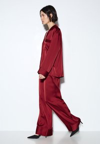 Sada vínově červených saténových pyžam s límečkem, zapínáním na knoflíky a kalhotami se širokými nohavicemi. Hladká textura, minimalistický design, s na míru ušitými manžetami a detaily na kapsách.
