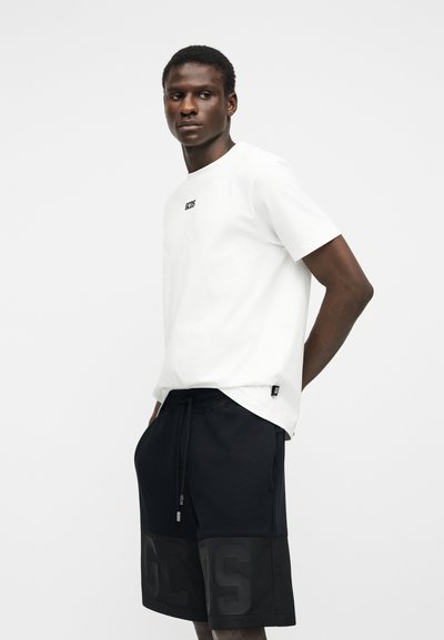 Άνθρωπος που φοράει λευκό πουκάμισο με κοντό μανίκι και μαύρο λογότυπο, καθώς και μαύρα σορτς με κορδόνι και μεγάλα ανάγλυφα γράμματα, στέκεται μπροστά σε απλό φόντο.