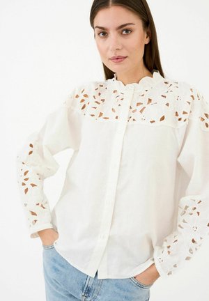 Žena nosi bielu košeľu na gombíky s kvetinovou čipkou na ramenách a rukávoch, v kombinácii so svetlomodrými džínsami, na bielom pozadí.