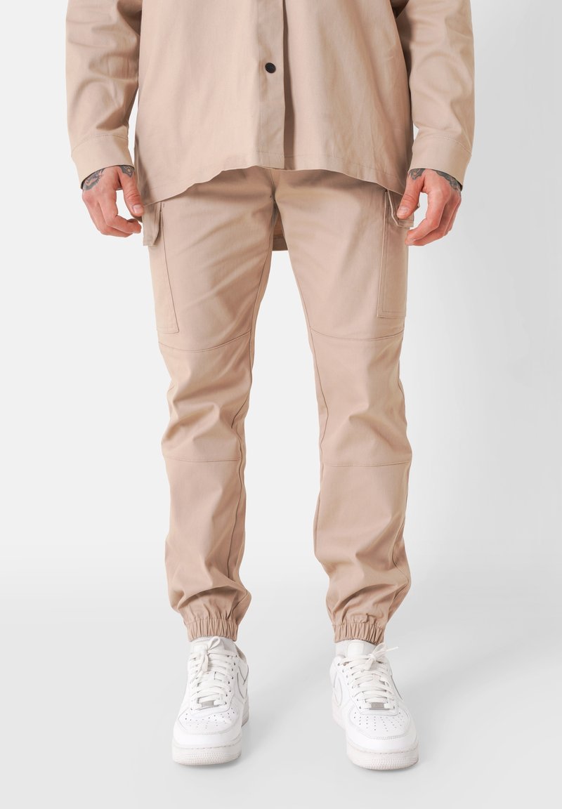 Homme portant un pantalon cargo beige à poignets élastiques, une chemise beige assortie et des baskets blanches, debout devant un fond uni.