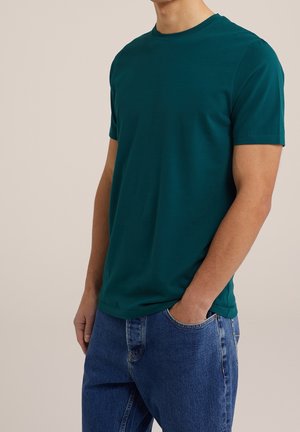 Teeppivihreä puuvillainen t-paita, jossa pyöreä pääntie ja lyhyet hihat, yhdistetty sinisiin farkkuihin. Paita on vartalonmyötäinen ja sileäpintainen.