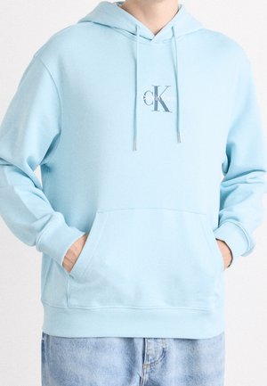 Osoba nosząca jasnoniebieską bluzę z kapturem Calvin Klein z przednią kieszenią i troczkami, w połączeniu z jasnoniebieskimi dżinsami.