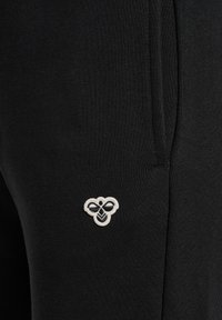 Sort stof med en lille hvid broderet bi-logo nær en lodret søm, del af et tøjs lommeområde.