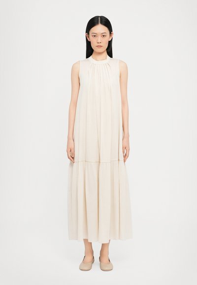 Robe sans manches beige avec un corsage plissé, une jupe fluide et un bas à étages. Texture lisse, longueur cheville, associée à des ballerines de couleur claire.