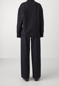 Persona che indossa una maglietta nera a maniche lunghe e pantaloni in denim larghi scuri, in piedi contro uno sfondo chiaro e uniforme, vista da dietro.