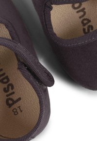 Chaussures en tissu violet foncé avec un extérieur doux, un bout rond et une sangle Velcro. L'intérieur présente une doublure brun clair avec le nom de la marque.