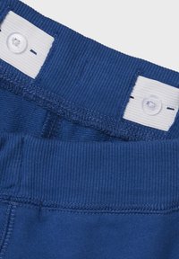 Ceinture en tissu bleu des pantalons, avec une texture côtelée et une étiquette blanche avec une fermeture à bouton. Les détails de couture sont visibles.