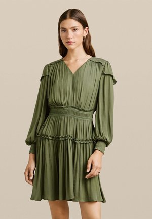 Vestido verde oliva con mangas largas abullonadas, escote en V, cintura fruncida y dobladillo con volantes. Hecho de un tejido suave y ligero con un diseño fluido.
