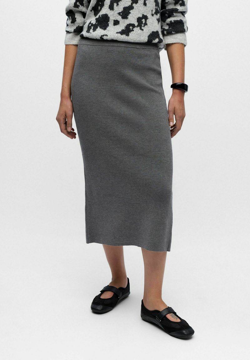 Jupe en tricot grise, longueur genoux, avec une texture lisse, dotée d'une taille ajustée. Associée à des chaussures noires à enfiler.
