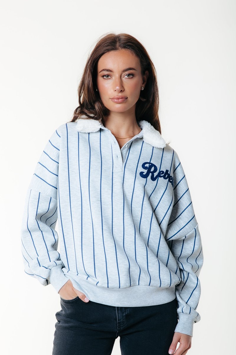 Világosszürke pulóver sötétkék függőleges csíkokkal, puha fehér gallérral, és sötétkékkel hímzett "Rebel" felirattal. Lazább szabású, bordás mandzsettákkal rendelkezik.