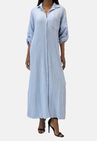 Robe bleu clair à manches longues et boutonnée, en tissu texturé, avec une poche poitrine ; présente des manches retroussées et descend en dessous des chevilles.