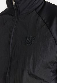 Svart zip-up jacka med en texturerad tygkonstruktion som ger en rynkad look och släta accenter. Broderad logo på bröstet.