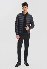 Kurtka puchowa w ciemnoszarym kolorze, pikowana struktura, zamek błyskawiczny z przodu, zestawiona z czarną koszulą i czarnymi spodniami. Model nosi czarne trampki.