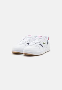 Hvite joggesko med en glatt læroverdel, kontrasterende marineblå og røde detaljer, perforerte elementer og såle i gummigummi. Lacoste-merke er synlig.