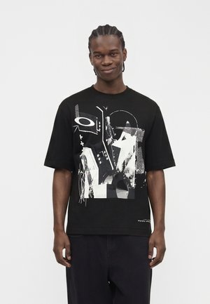 Mand med flettet hår iført en sort oversized t-shirt med hvidt abstrakt grafisk tryk og sorte bukser, stående mod en hvid baggrund.