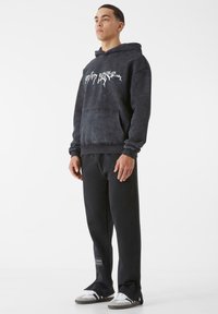 Juodas džemperis su priekinėmis kišenėmis ir baltu grafiniu tekstu, derinamas su juodomis sportinėmis kelnėmis, ant kurių yra logotipo aplikacija, vilkimas su žemais sportbačiais.