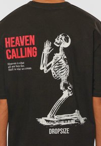 Sort t-shirt med en hvid skelet-illustration på ryggen, med rødt tekst der siger "HEAVEN CALLING" og et citat nedenfor.