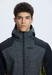 Giacca con zip in un tessuto grigio texturizzato, spalle nere e dettagli verdi. Presenta un cappuccio e un piccolo logo sul petto.