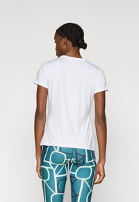 Λευκό αθλητικό τοπ με κοντά μανίκια και στρογγυλή λαιμόκοψη, συνδυασμένο με πετρόλ κολάν που φέρει γεωμετρικό μοτίβο σε μπλε και κρεμ αποχρώσεις.