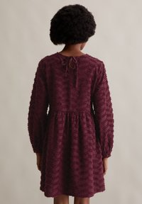 Bordeauxfarbenes Kleid mit langen Ärmeln, strukturierter Stoff mit erhabenen Zickzackmustern, elastischem Taillenbund und einer Bindeschließe am Rücken.