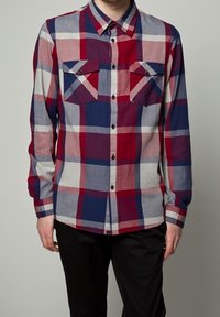 Man i långärmad skjorta med röd, blå, vit och rosa rutmönster och svarta byxor.