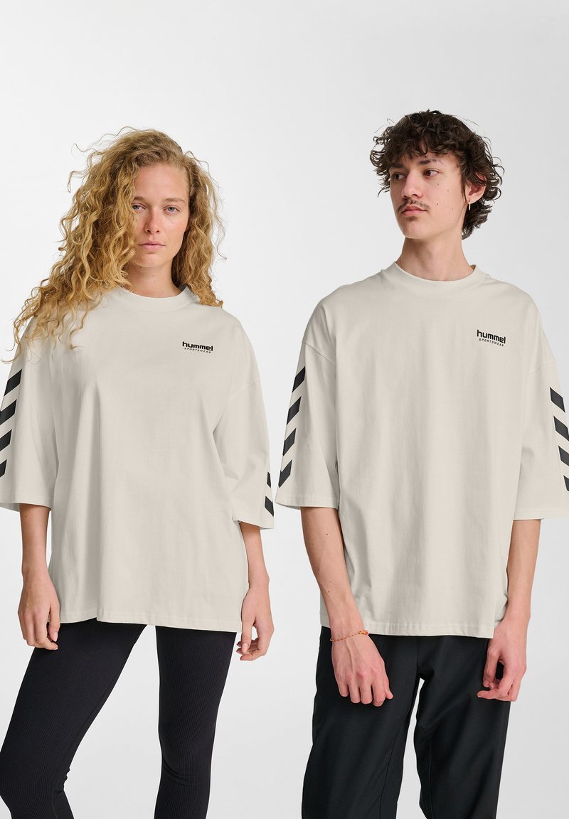 Licht beige oversized t-shirt met zwarte gestreepte accenten op de mouwen en een klein logo op de borst. Katoenen materiaal, relaxte pasvorm.