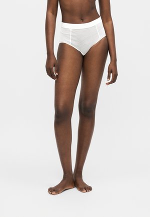 Kvinde iført hvide højtaljede trusser med lodrette striber, stående barfodet mod en ensfarvet lys baggrund.