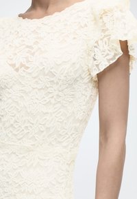 Közelkép egy krém színű csipkeruháról, amely virágmintás és fodros rövid ujjas, a személy felső törzsén és karján látható.