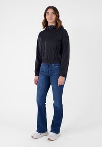 Giacca nera corta con zip e tasche laterali, abbinata a jeans a zampa d'elefante blu e sneakers bianche. Tessuto morbido e vestibilità casual.