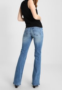 Nainen seisoo vaaleansinisissä levitemissingissä ja mustassa hihattomassa paidassa, kuvattuna selkäpuolelta yksinkertaista taustaa vasten.