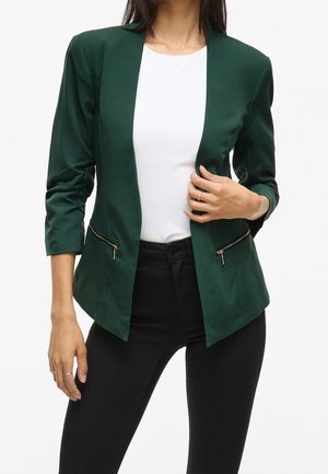 Woman wearing dark green blazer with zipper pockets, white top, and black pants, posing with one hand on blazer and the other relaxed.