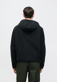 Czarna kurtka z kapturem wykonana z gładkiego materiału, o luźnym kroju i elastycznych mankietach, noszona z luźnymi oliwkowozielonymi spodniami.