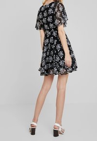 Mustat kukalliset mekko, jossa on läpikuultavat liehukeet ja liehuva helma, sekä valkoisia kasvitulostuksia. Ruskeat paksukorkoiset sandaalit täydentävät asun.