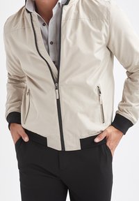 Beige leichte Bomberjacke mit durchgehendem Reißverschluss vorne, seitlichen Reißverschlüssen, schwarzen gerippten Bündchen und Saum, mit glatter Textur und minimalistischem Design.