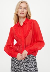 Rote durchsichtige Bluse mit rüschem Ausschnitt, Knöpfen auf der Vorderseite und langen Ärmeln. Kombiniert mit einem gemusterten schwarz-weißen Rock.