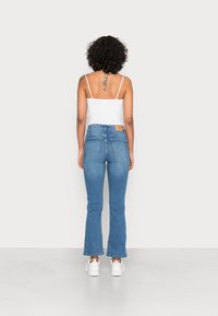 Calças de ganga azul em flare com cintura alta e bolsos traseiros, combinadas com uma blusa branca de alças finas e ténis brancos.