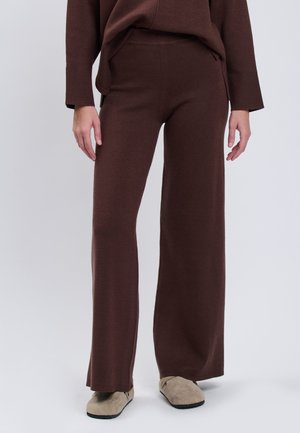 Person iført brune strikkede vidde bukser og matchende sweater, kombineret med beige ruskind slip-on sko, stående på lysegråt gulv.