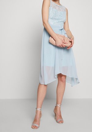 Femme portant une robe sans manches en dentelle bleu clair avec un ourlet asymétrique, des sandales à talons hauts argentées, tenant une pochette rose pâle.