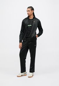 Jaunas vyras su pintomis plaukų sruogomis, vilkintis juodą Reebok treniruočių kostiumą ir baltus sportinius batus, stovi su viena ranka kišenėje ir žiūri į šoną.